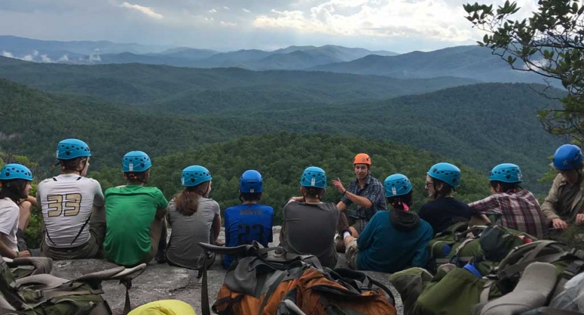 A group of students wearing helmets sit in a line and listen to an instructor speak. In the background, there is a vast and green mountainous landscape.